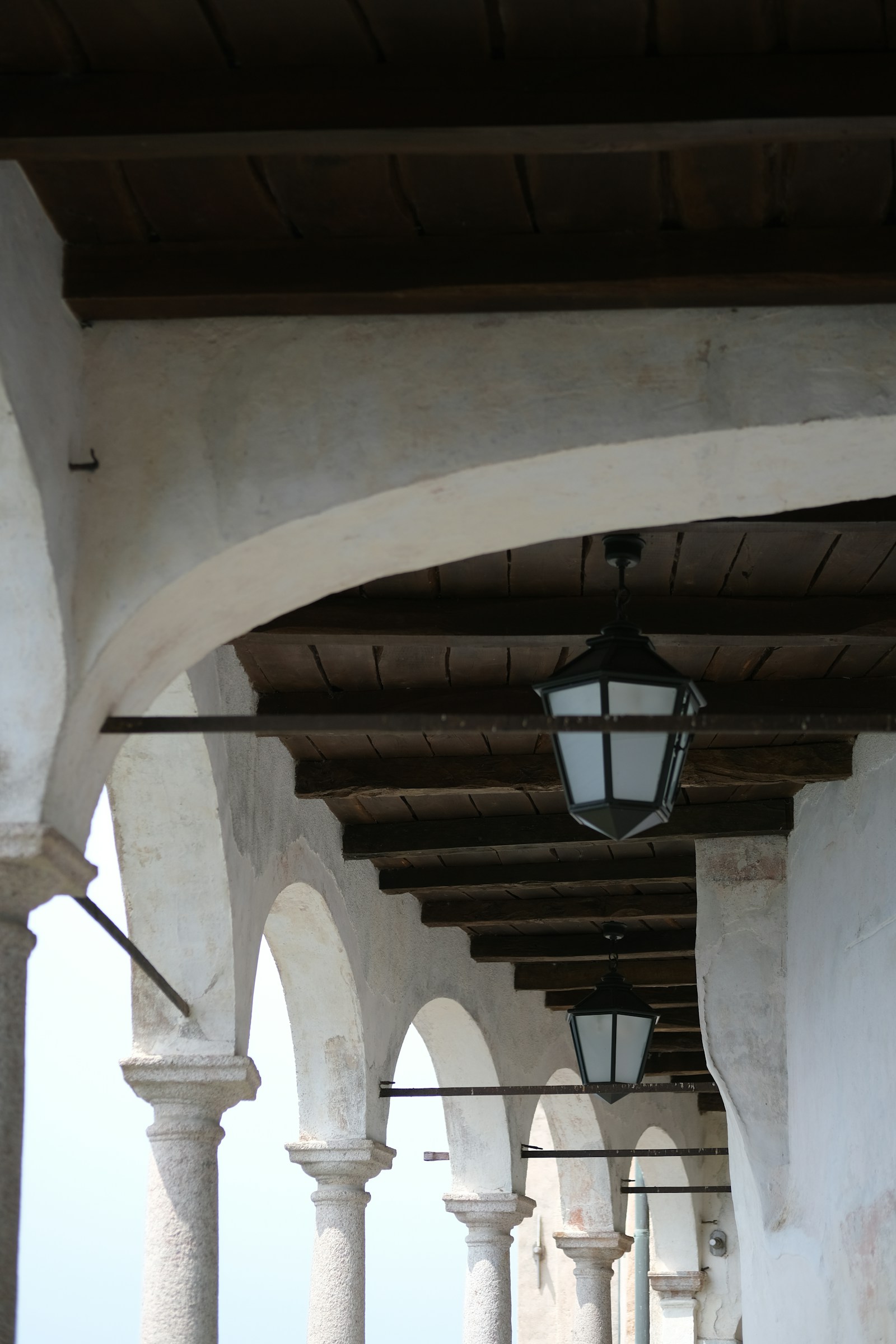 Warm arcade corridor of whitewashed stone arches with dark beamed ceilings and hanging lanterns, backlit by soft light