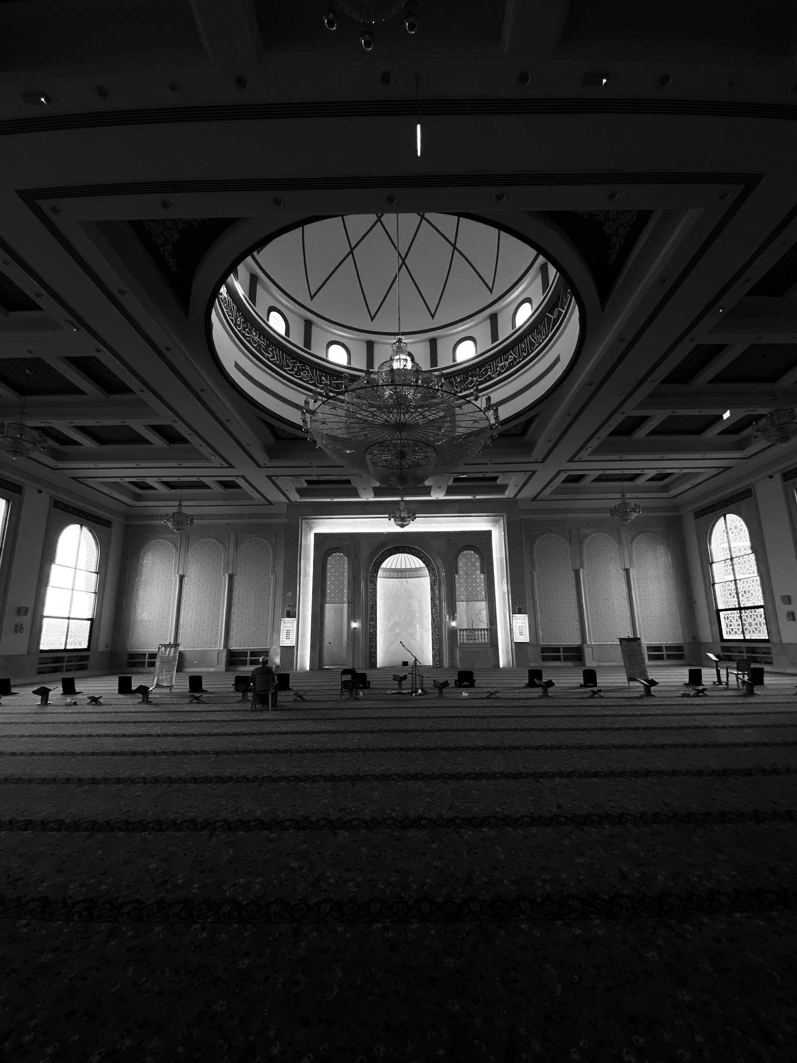 Dim mosque prayer hall beneath a patterned dome, lit by a chandelier and arched windows